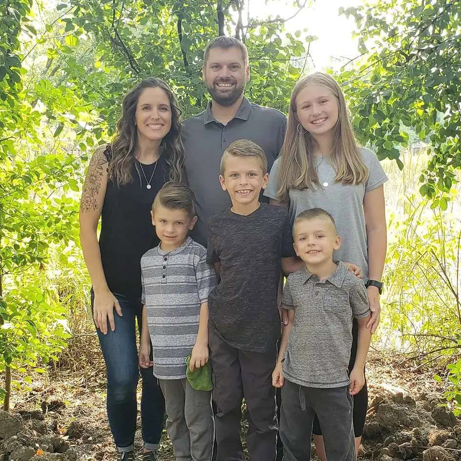 )Sarah, her husband Andy and their four kids smile while standing next to a tree.