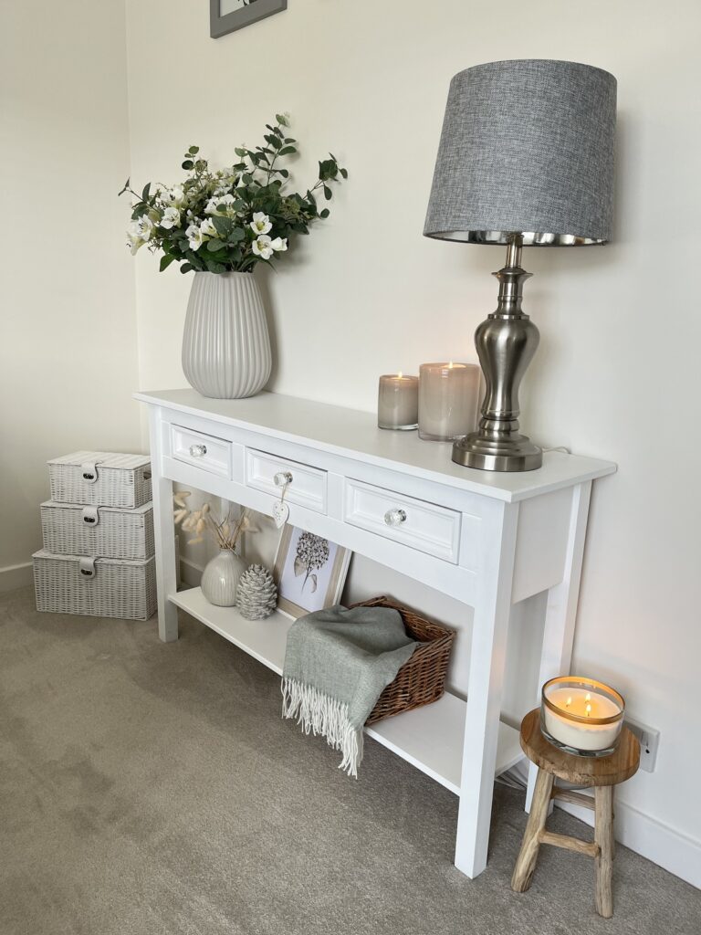 A white console table with a lamp, flowers and blankets.