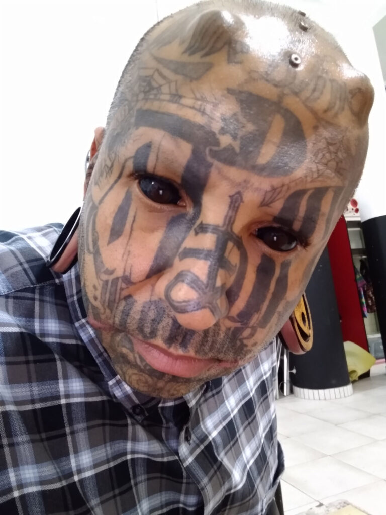 Renato with black eyes, tattoos and horns while wearing a checkered shirt. 
