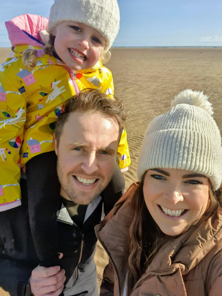 Chris smiling with his wife, Jo and daughter, Millie while on a beach.