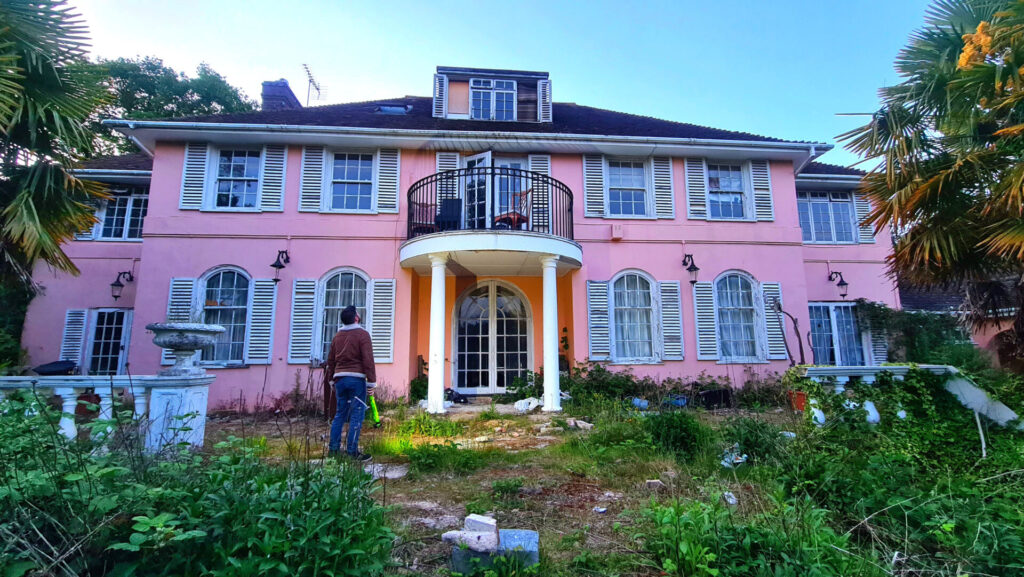 A pink mansion with multiple windows and white pillars sits by an unkept piece of lawn