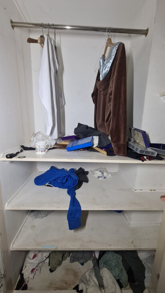 A white shirt and brown jumper still hang from the wardrobe with a blue garment underneath on the shelves. 