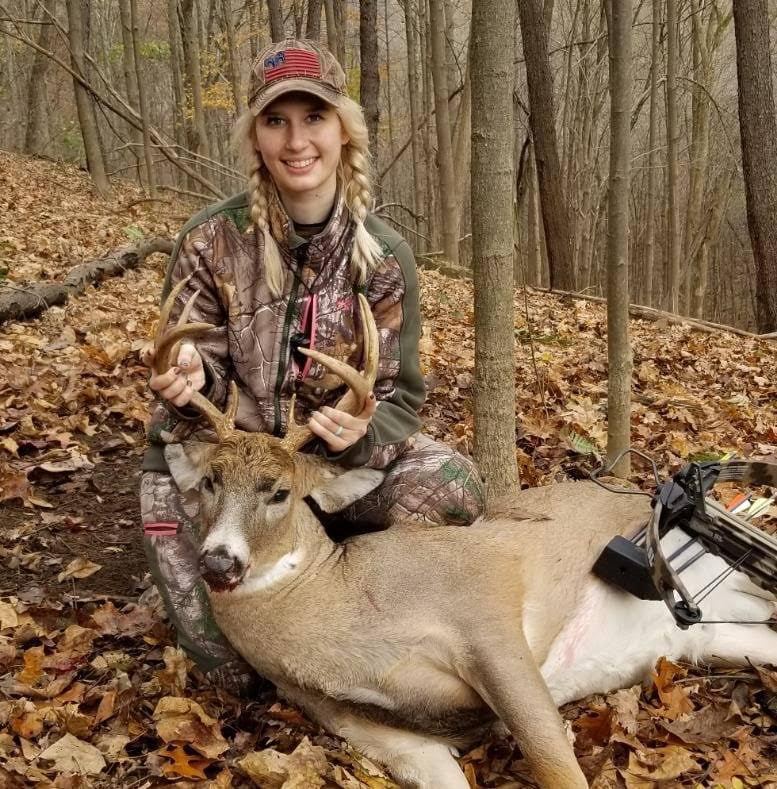 Katelyn smiles in camp gear while holding up a dead buck by its antlers. 