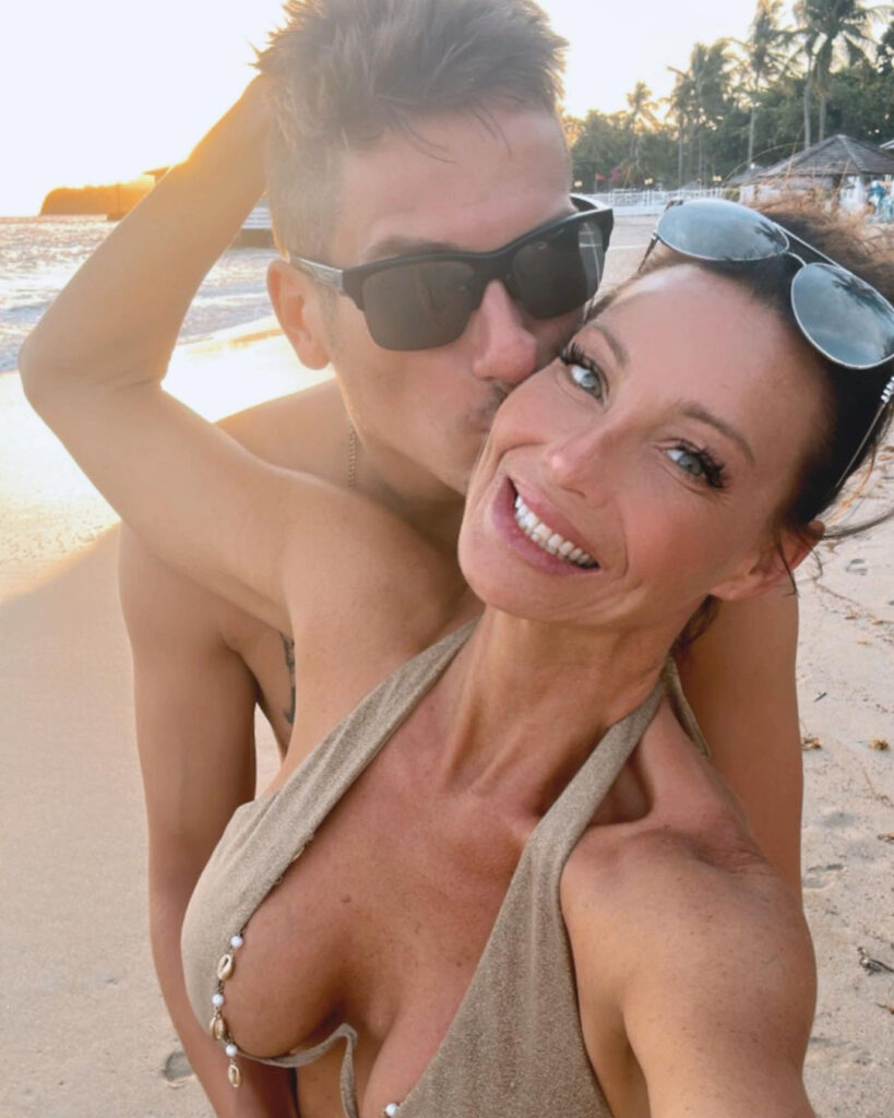 Sarah poses in a bikini while on a beach holding Trenton's head who is kissing her.